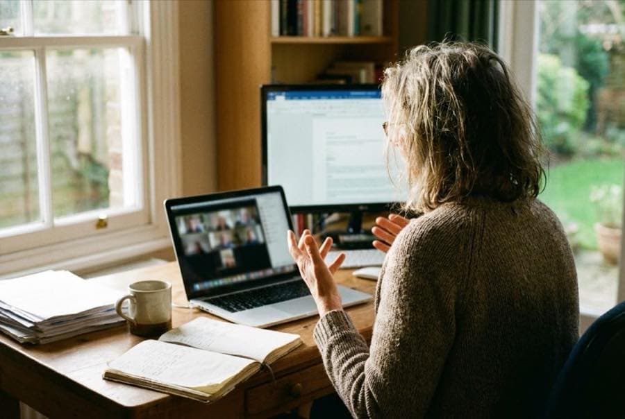 An adviser at a desk at home, on a video call, with papers and a notebook open beside the screen.