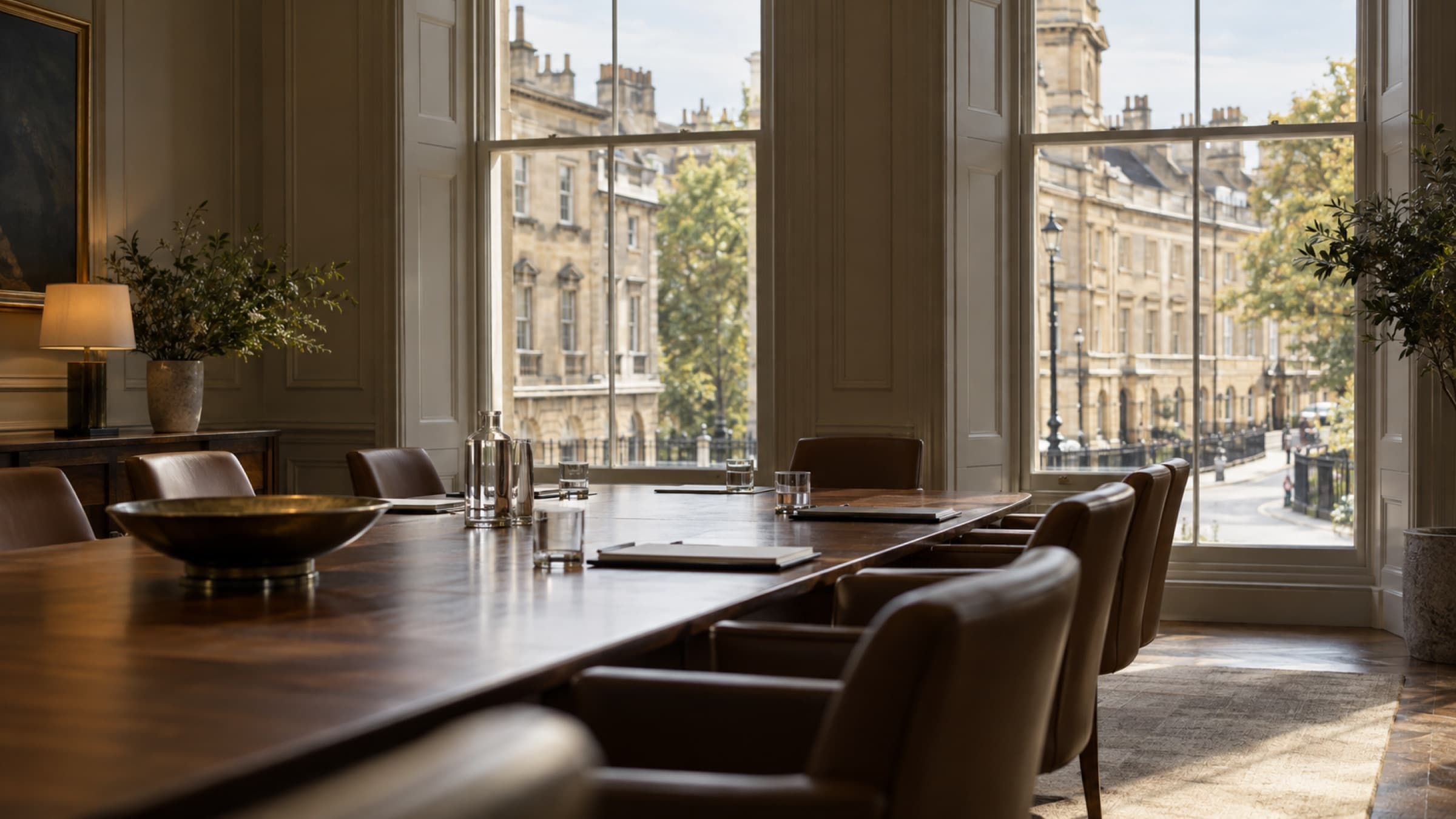 An empty meeting room with a wooden table and leather chairs, overlooking a street of stone buildings through tall windows.