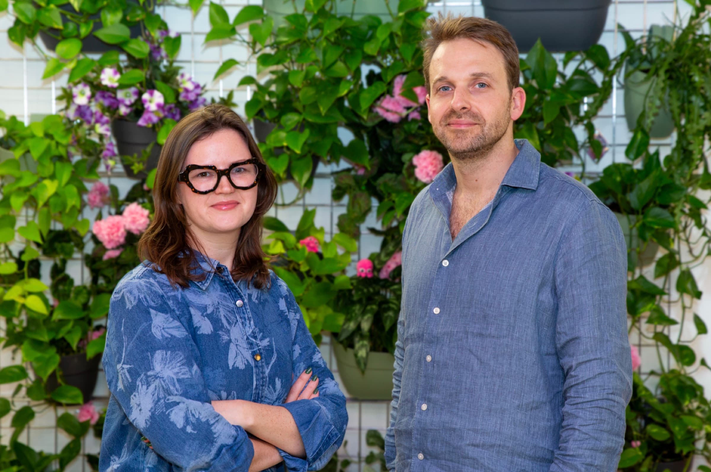 Two people in blue shirts standing in front of a planted wall with pink and purple flowers.