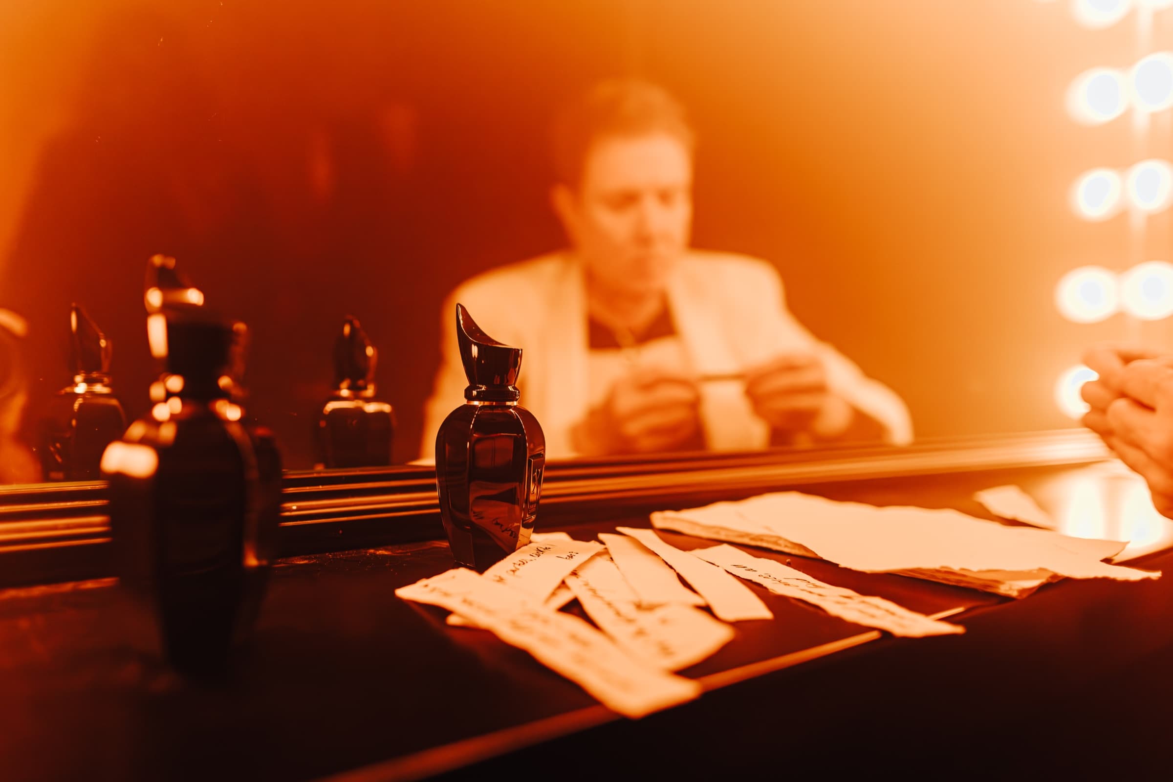 Perfume bottles and paper scent strips on a dressing table, with a person reflected in a bulb-lit mirror in warm orange light.